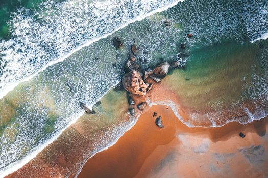 Drone view of foaming waving sea with colorful water and sandy coast with big boulders