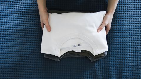 Hands folding a white shirt on a blue textured bedspread, focusing on organizing clothes.