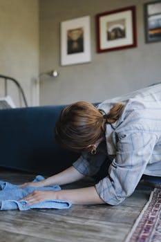 A woman in a striped shirt wipes the wooden floor of a bedroom, emphasizing housekeeping.