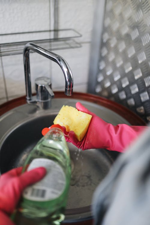 Free Hands wearing rubber gloves pouring detergent on a sponge over a kitchen sink. Stock Photo