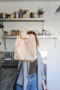 Close-up of a person holding a brown paper bag labeled 'Delivery' in a kitchen setting.