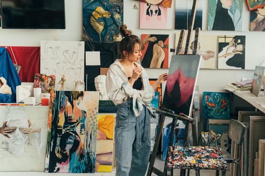 A woman artist in her studio thoughtfully gazes at her canvas amidst a collection of her artwork.