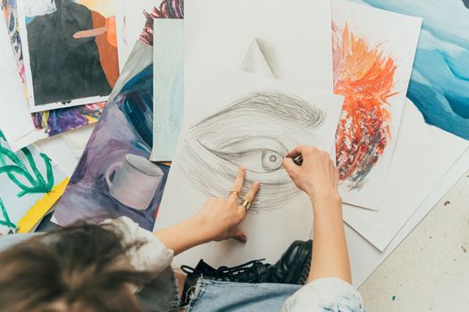 An artist drawing an eye while surrounded by various colorful artworks, using pastels indoors.
