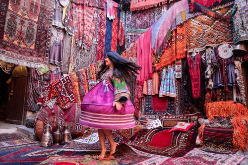 Free A woman in a vibrant pink dress spins joyfully among colorful Turkish carpets at a traditional market. Stock Photo