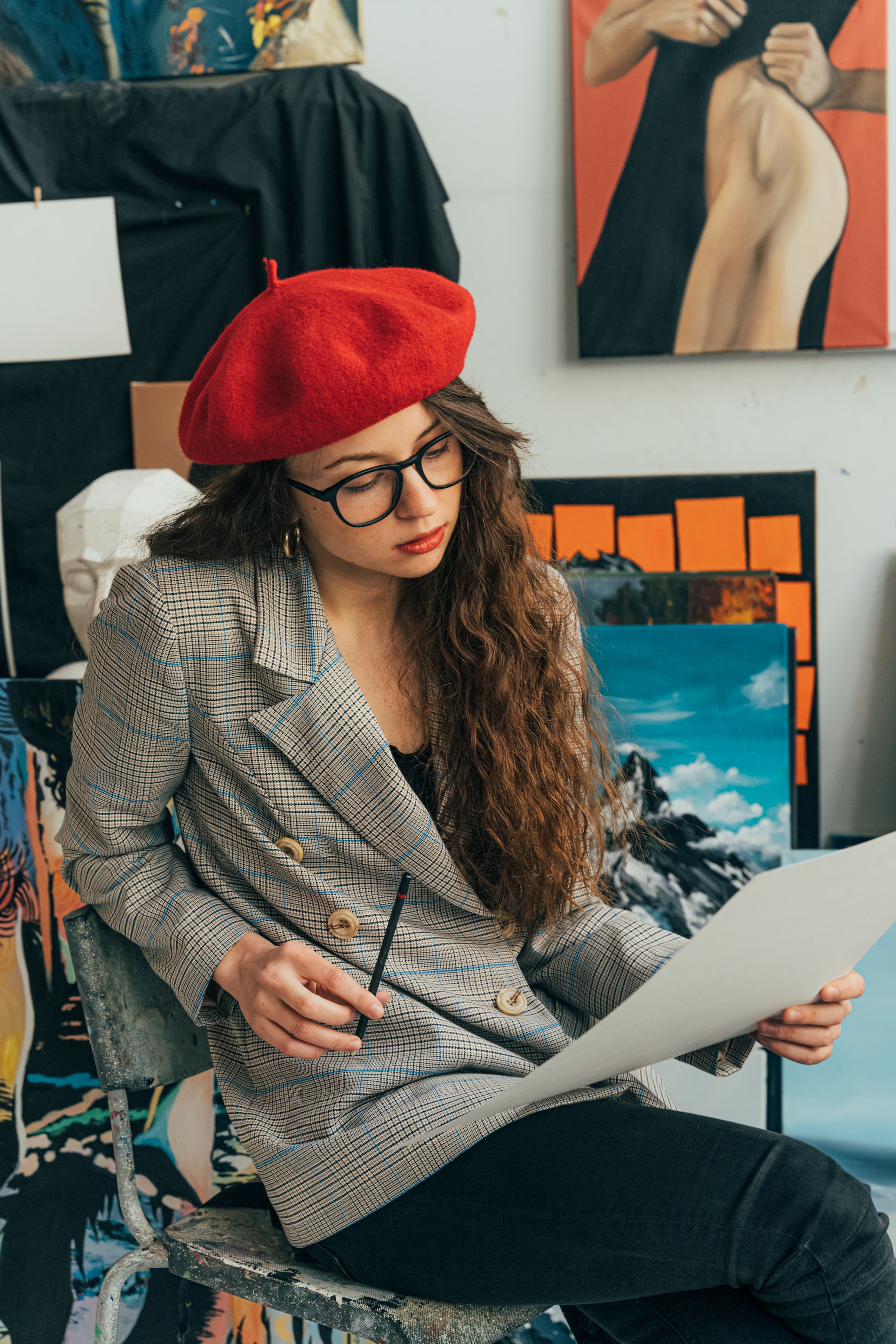 Young woman artist wearing a red beret sitting in studio analyzing artwork.