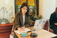咖啡馆里，一位年轻女子正对着笔记本电脑写笔记，桌上放着咖啡杯和壶
