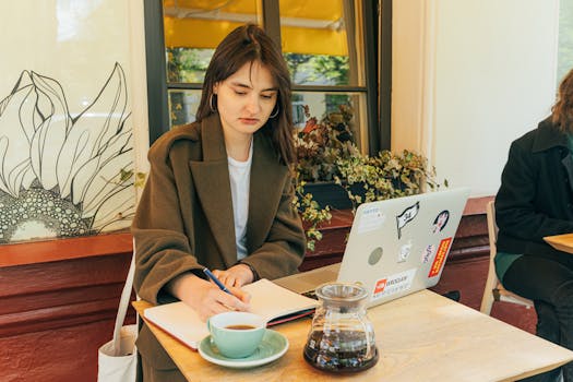 Young woman working at café, writing notes beside laptop with coffee cup and pot on table.