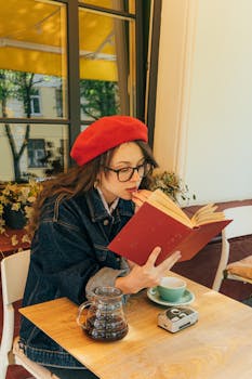 Woman wearing a red beret reads a book in a cozy cafe setting, sipping coffee.