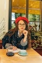 一位戴着红色贝雷帽和眼镜的时尚女性，正坐在咖啡桌旁，手里拿着咖啡和笔记本