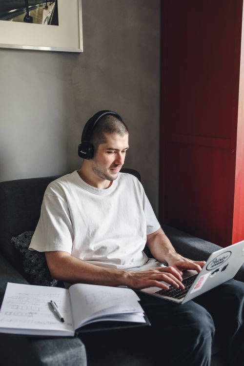 Free Young man sitting indoors, wearing headphones, and typing on a laptop for remote work. Stock Photo