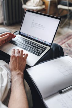 Close-up of a person using a laptop with a notebook open beside it, focusing on hands and typing.