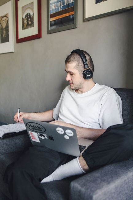 Adult man sitting on a couch, writing in a notebook while using a laptop, wearing headphones.