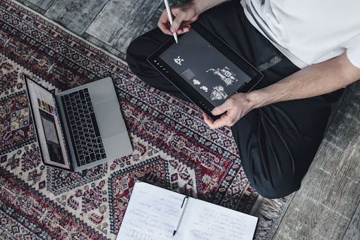 A person draws on a tablet while sitting on a carpet with a laptop and notebook beside them.