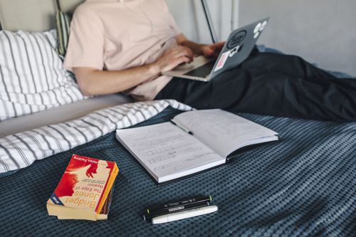 Free Young adult sitting on bed using laptop with books and notes spread around, creating a cozy study environment. Stock Photo