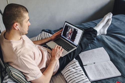 Free Young man on bed video calling using laptop and earphones, surrounded by notes. Stock Photo