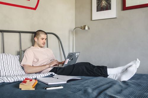 Free Man working on laptop from bed in modern bedroom. Comfortable remote work setup with a relaxed vibe. Stock Photo