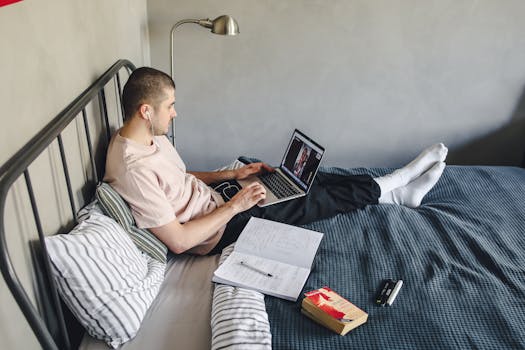 A man sitting on a bed with a laptop, engaged in a video call, indoors.