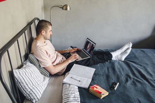 Free A man sitting on a bed with a laptop, engaged in a video call, indoors. Stock Photo