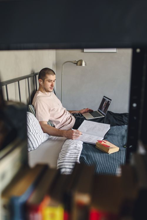 Free A man studying in bed with a laptop and books, illustrating remote learning. Stock Photo
