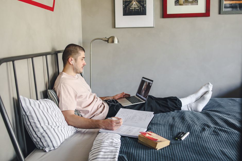 Young man studying online from home, comfortably seated on a bed with laptop and books.