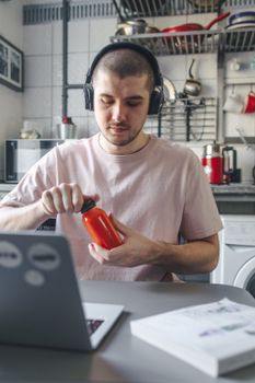 A young man in a kitchen using a laptop while wearing headphones. Engaged in multitasking.