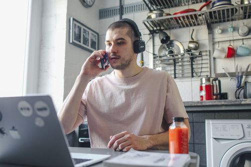 Free Adult man multitasking at home with laptop and phone, comfortable setup. Stock Photo