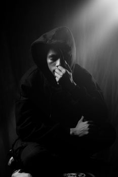 一个穿着黑色连帽衫、神情悲伤的男子闭眼坐着，用兜帽遮住了脸