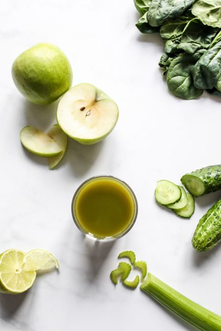 A tall glass of pale green juice made from cucumber, apple, kale, and spinach, with a lemon half and a whole green apple sitting beside it on a wood counter.