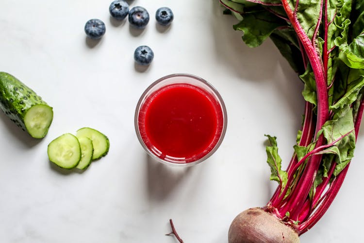 Red Juice In Clear Drinking Glass, Beetroot, Sliced Cucumber And Blueberries On White Table