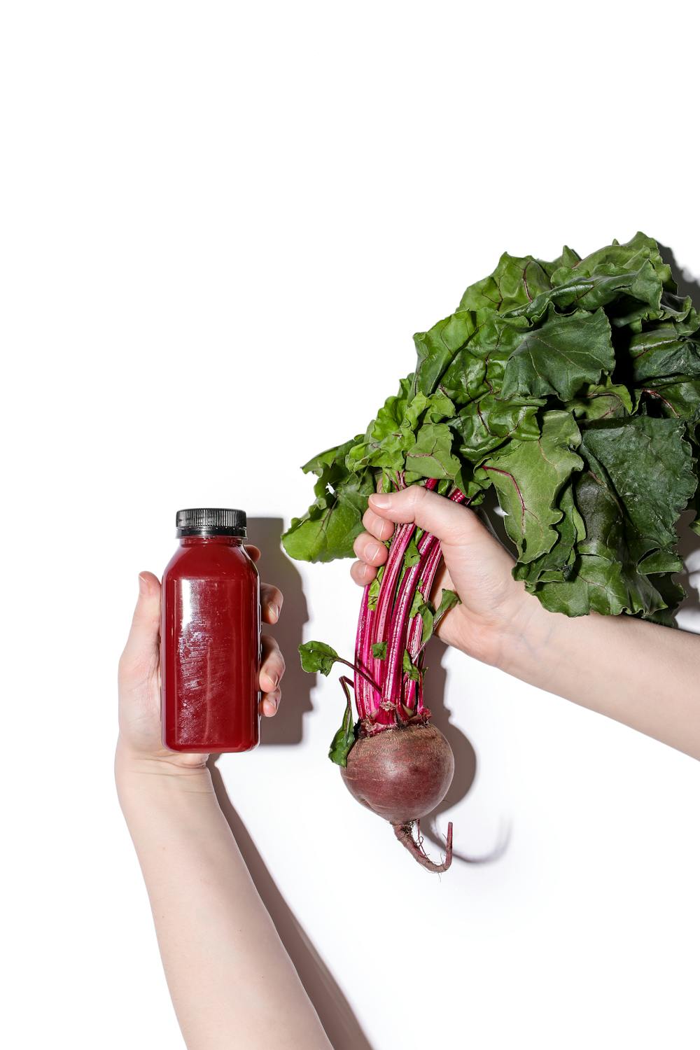 white person holding beet juice in one hand and fresh beets from the stem in the other hand on white backdrop