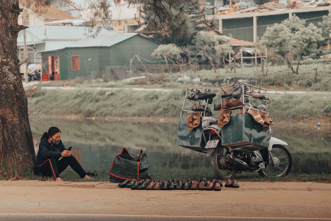 Full body of focused female street seller in casual clothing and face mask surfing internet on cellphone while selling shoes by road next to pond in settlement