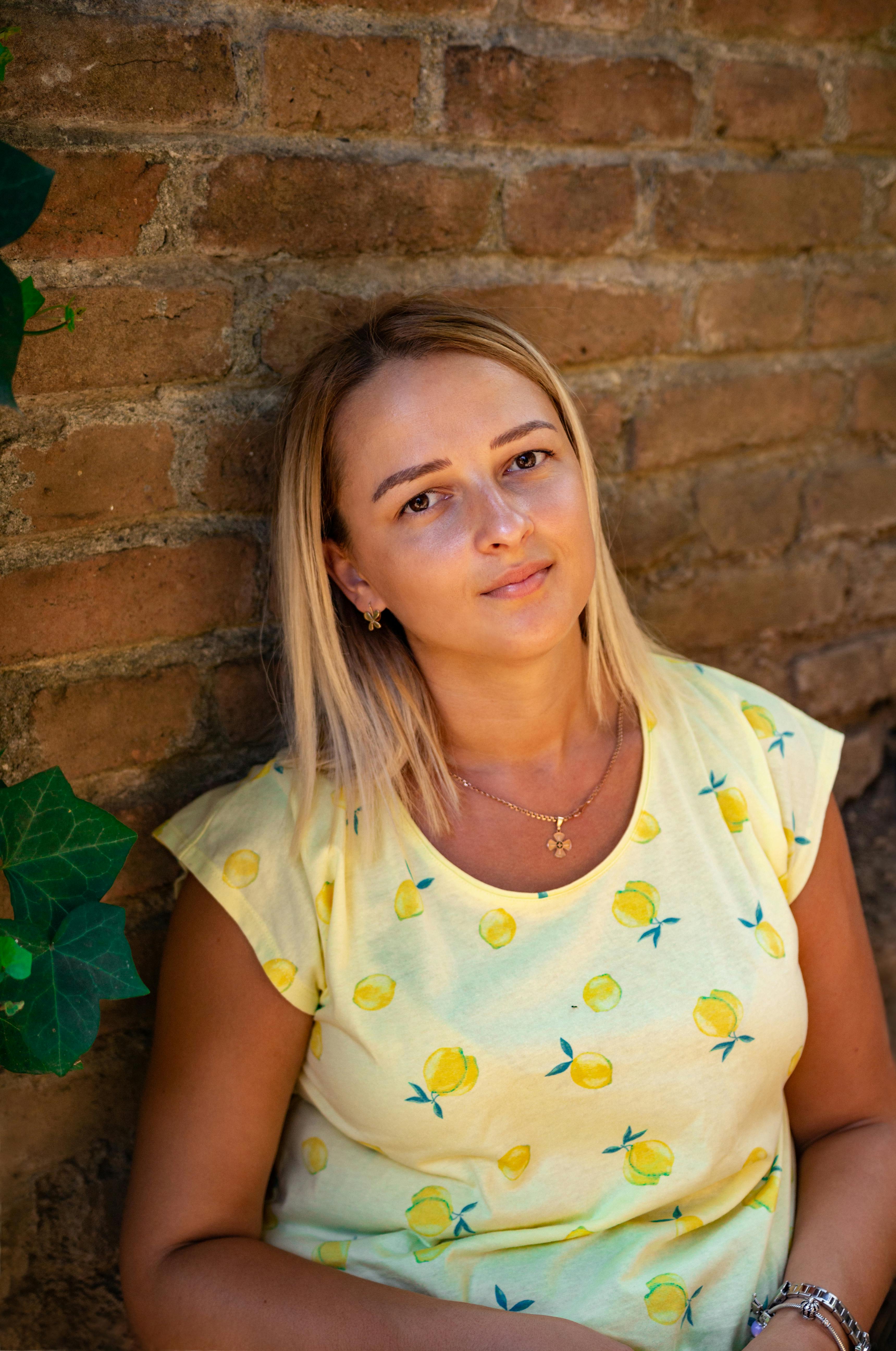 A young woman wearing a yellow lemon print shirt poses against a rustic brick wall.