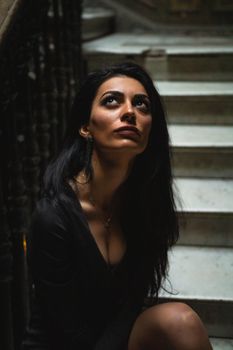 Artistic shot of a woman sitting on a vintage staircase, looking upward in a moody, dimly lit interior.