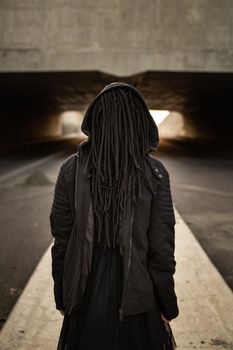 一个留着辫子、身穿连帽夹克的人站在一条阴森的城市隧道中，营造出神秘的氛围