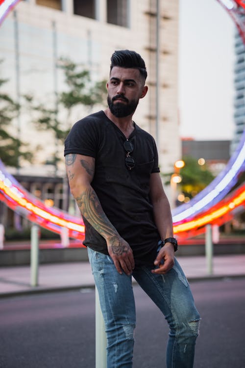 Confident bearded ethnic male with tattoos in casual clothes leaning on metal pole in urban area while looking away and contemplating
