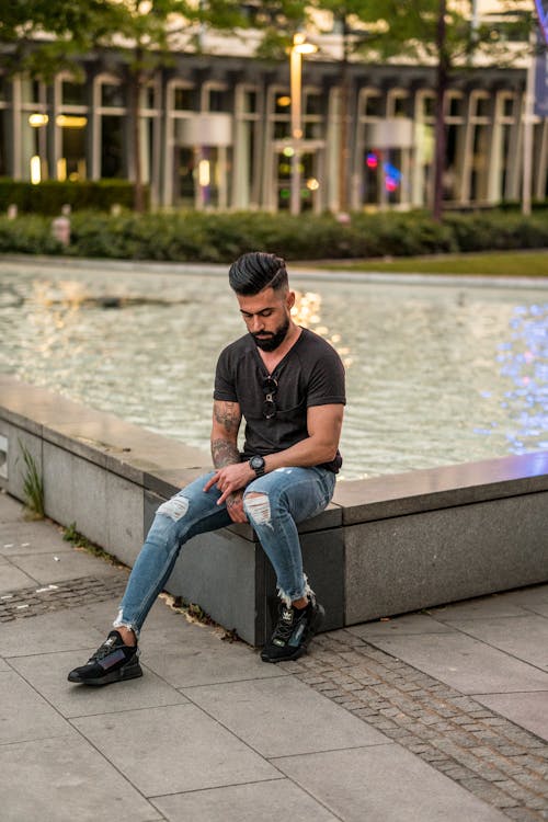 Full length of bearded ethnic man in casual outfit sitting on stone border and thinking while looking down in urban area