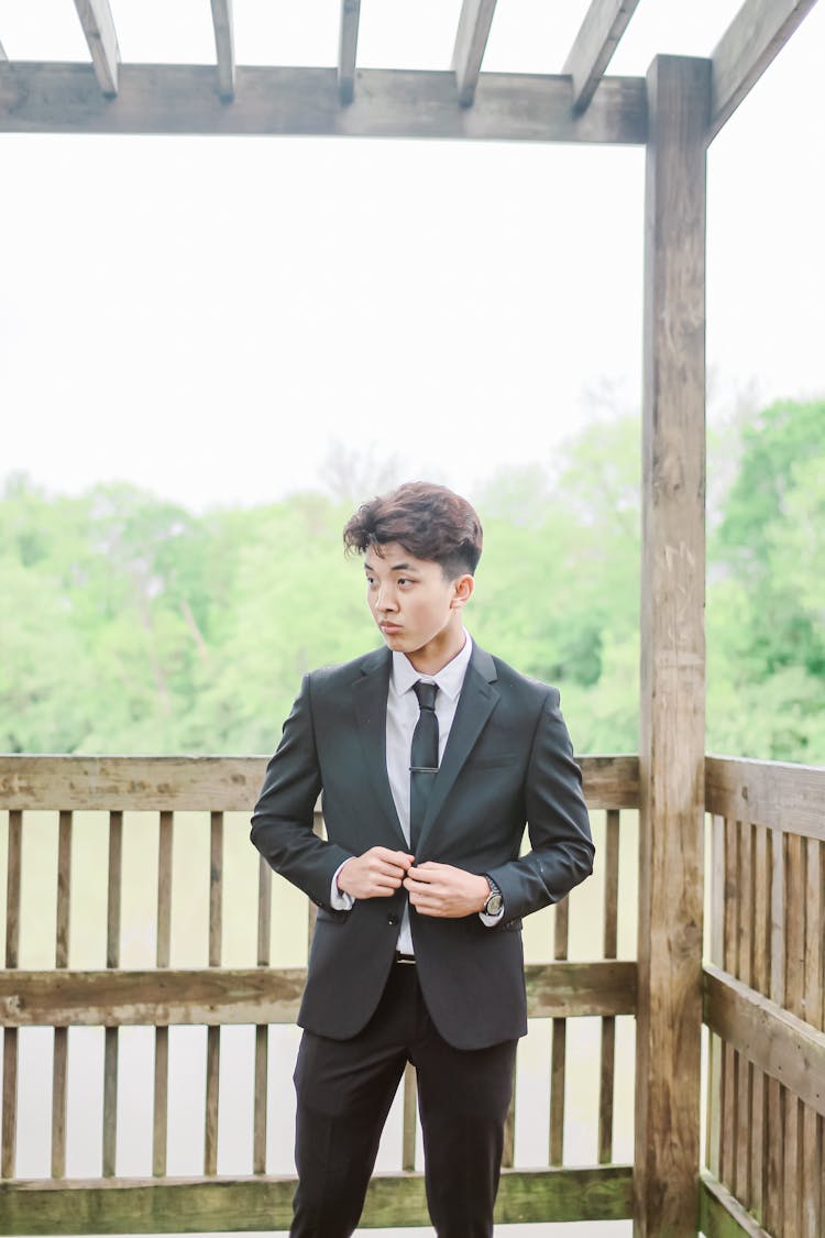 Man In Black Suit Jacket And Black Pants Standing Near Brown Wooden Fence