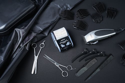 Overhead view of cash register surrounded by comb and metallic hairdressing scissors composing with clipper and hairpins on black cloth in bright room