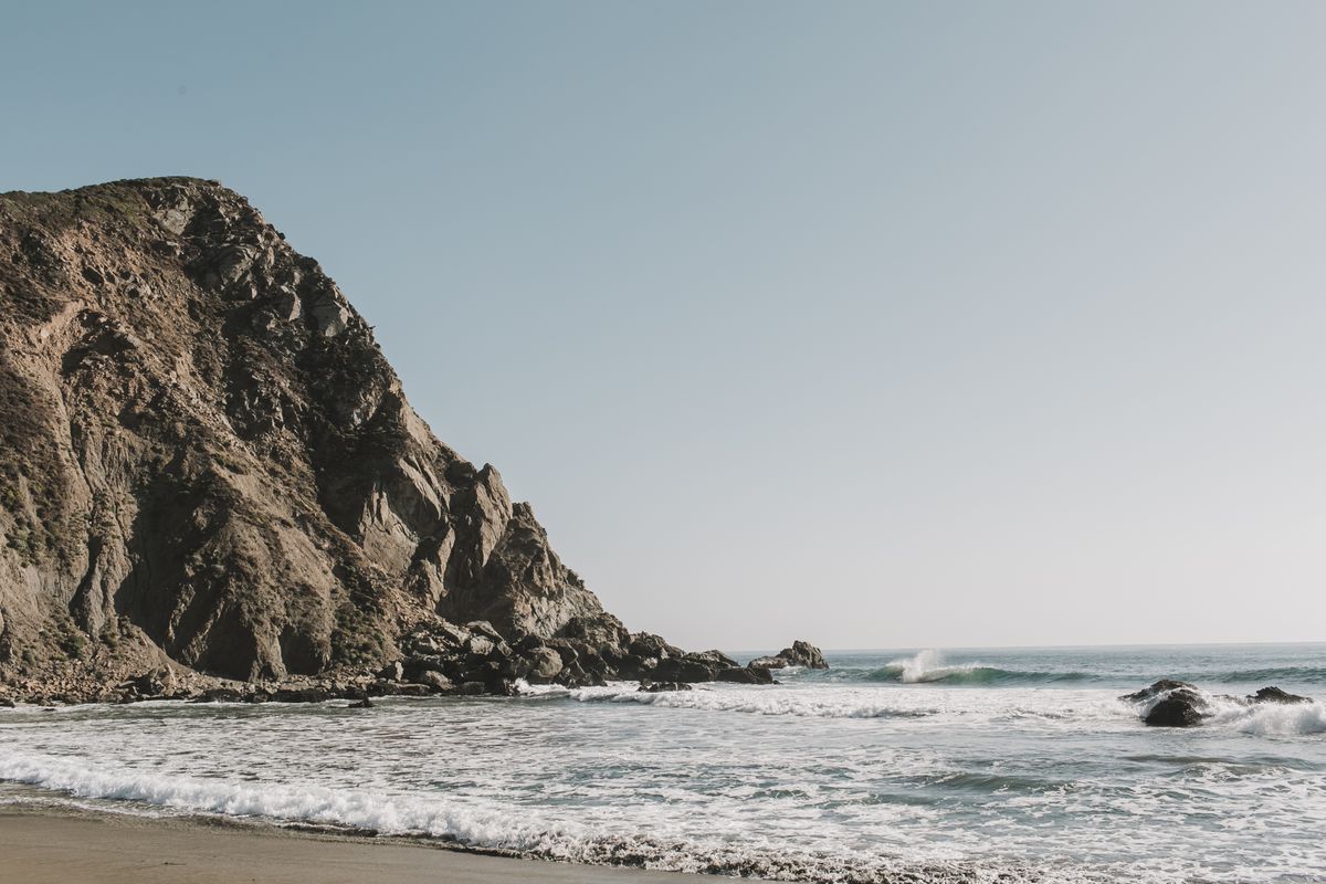 A Big Sur beach cove with a dark craggy headland on the left, white surf rolling onto the sand, and a small wave breaking offshore under a pale sky.