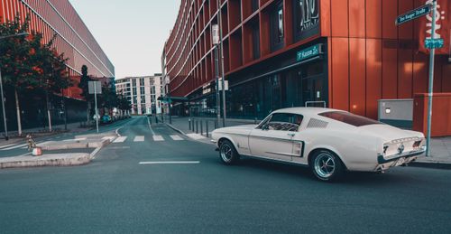 Free A classic vintage car driving through a modern urban street corner with orange buildings. Stock Photo