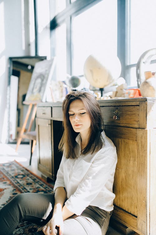 Free Woman in White Long Sleeve Shirt Sitting Near Wooden Cabinet Stock Photo