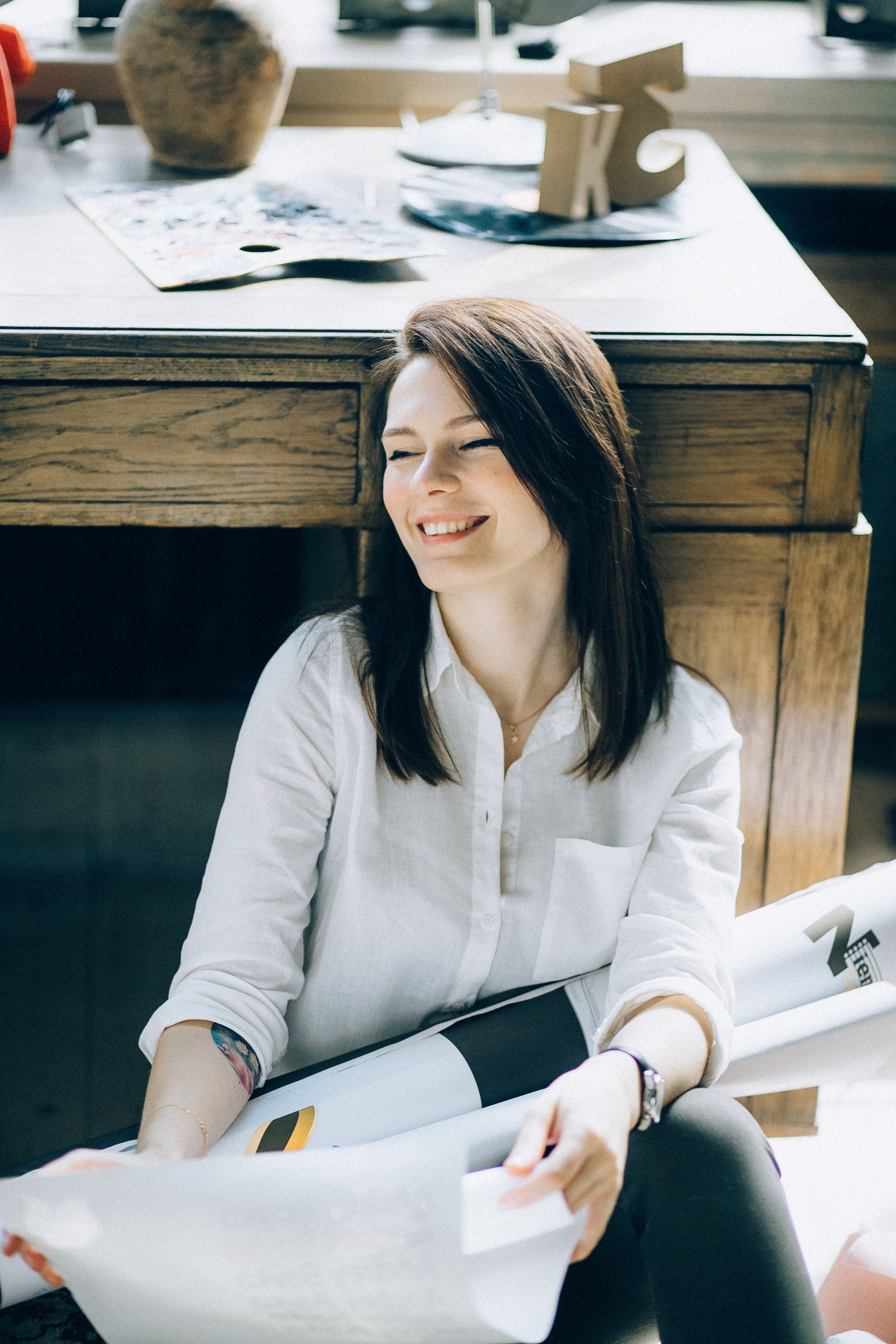 Relaxed woman sits under a wooden desk holding papers, smiling in a bright indoor setting.