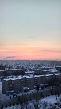 From above of old low cost grey concrete paneled five srorey house surrounded by paved road and settlement under light blue pink evening sky with sun on horizon