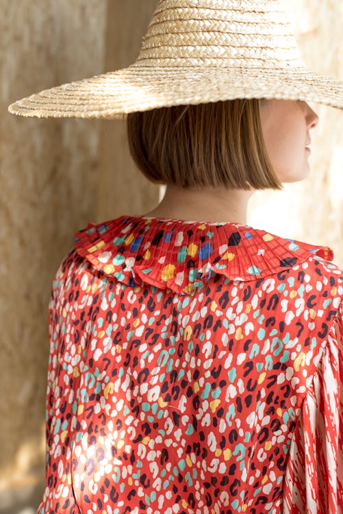 Free Back view unrecognizable stylish lady in colorful red summer dress and straw hat with short blond hair standing near beige wall and looking away Stock Photo