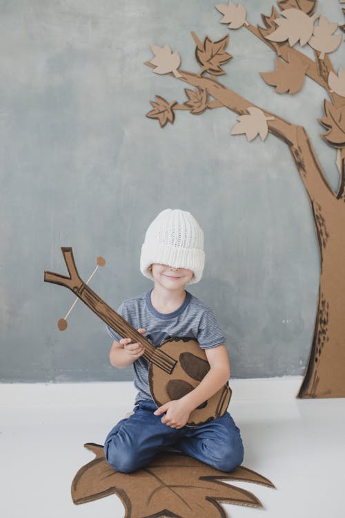 Full body smiling cute little boy in casual clothes with eyes covered with white knitted hat sitting on floor with toy dombra against gray wall and carton fake tree