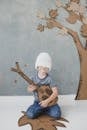 一个微笑可爱的小男孩，穿着休闲服，戴着白色针织帽，坐在地板上，玩具圆顶靠着灰色的墙壁和纸箱假树