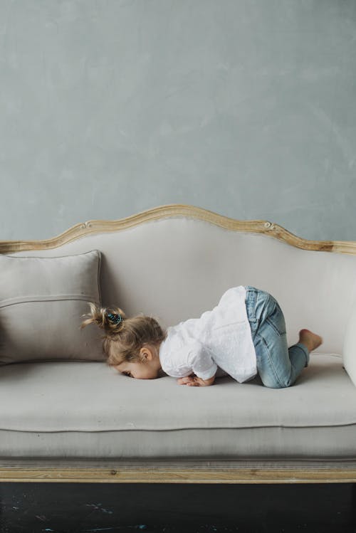 Side view full body adorable little girl in casual clothes lying on cozy comfortable sofa with face down against blue wall on daylight