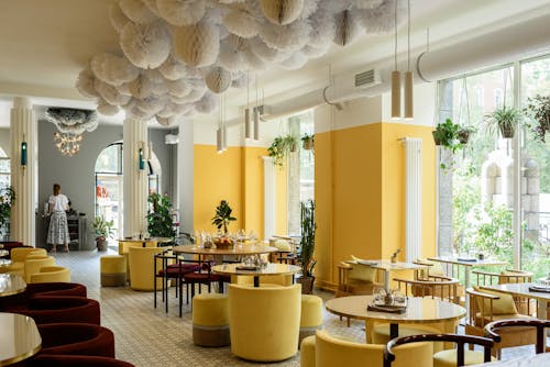 Creative interior design of modern spacious cafe with bright yellow walls and armchairs decorated with green plants and cozy lamps with big windows against lush greenery