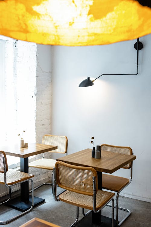 Cozy interior design of contemporary creative cafe with simple wooden tables and chairs under big yellow lamp placed against light blue wall