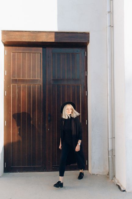 A fashionable woman poses elegantly against a rustic wooden door in bright daylight.